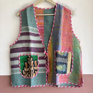 May include: A sleeveless waistcoat featuring a patchwork design with various colourful patterns, including stripes and floral motifs. The garment has two front pockets, a tie closure at the centre, and is finished with a pink and white striped trim.