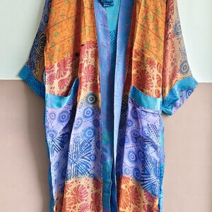 May include: A patchwork kimono robe in shades of orange, blue, and purple. The robe features floral and geometric patterns, with a blue trim and two front pockets. The garment is hanging on a wooden hanger.
