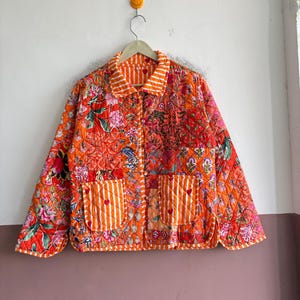 May include: A quilted jacket featuring a vibrant orange base with a multi-coloured floral pattern. The garment includes a collar and cuffs with white and orange stripes, two front pockets with white stripes and small red hearts, and a button closure.