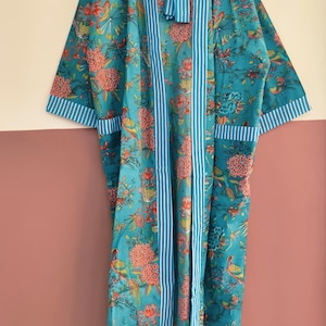 May include: A long-sleeved teal robe featuring a vibrant floral and bird pattern in shades of orange, pink, and green. The garment includes a white and blue vertical striped trim along the front opening, sleeve cuffs, and two front pockets.