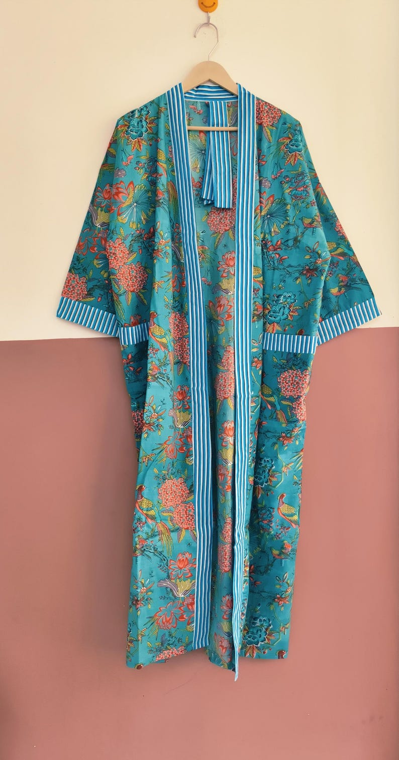 May include: A long-sleeved teal robe featuring a vibrant floral and bird pattern in shades of orange, pink, and green. The garment includes a white and blue vertical striped trim along the front opening, sleeve cuffs, and two front pockets.