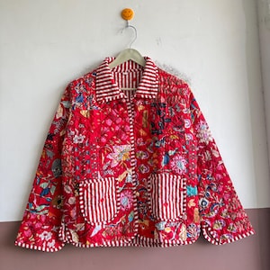 May include: A quilted jacket in a vibrant red hue featuring a multi-coloured floral pattern throughout. The garment includes a collar, cuffs, and two front patch pockets with a white and red vertical stripe design, finished with a button closure.