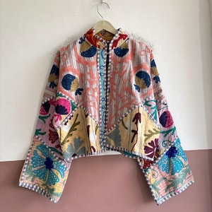 以下が含まれることがあります： ピンク、ブルー、イエロー、ホワイトの色合いで複雑な花柄の刺繍が施されたパッチワークデザインのクロップドジャケット。長くて幅の広い袖、マンダリンカラー、全体にステッチが見えるフロントオープニングが特徴です。