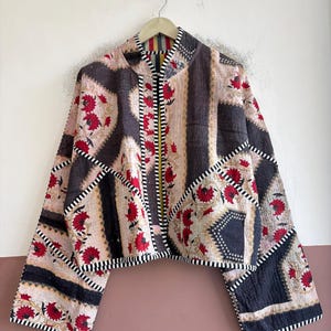 May include: A cropped jacket featuring a patchwork design with black, cream, and red floral patterns. The garment has long, wide sleeves, a stand-up collar, and black and white striped trim along the edges and seams throughout the piece.