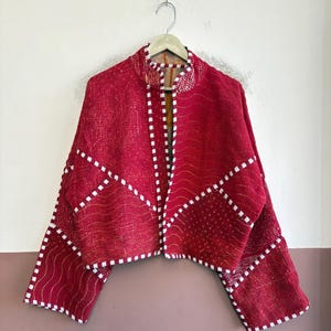 Könnte beinhalten: Eine kurz geschnittene Jacke in leuchtendem Magentarot mit Stehkragen und weiten Ärmeln. Das Kleidungsstück besteht aus gesteppten Stoffbahnen, die an den Kanten, Nähten und Ärmelabschlüssen mit einem weiß-roten Karomuster verziert sind.