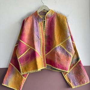 May include: A cropped jacket featuring a quilted texture with a vibrant tie-dye pattern in shades of pink, orange, and yellow. The garment has long sleeves, a mandarin collar, and is accented with white and yellow striped trim along the seams.