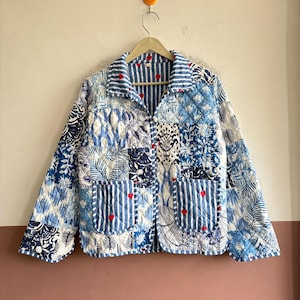 May include: A quilted jacket featuring a patchwork design of blue and white floral, botanical, and striped patterns. The garment includes a collared neckline, long sleeves, two front patch pockets, and small red heart embroidery throughout.