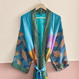 May include: A colorful, patterned robe with a tie closure, hanging on a wooden hanger. The robe features a mix of blue, green, yellow, and purple fabrics with leaf and abstract designs. The robe is made of a lightweight material.