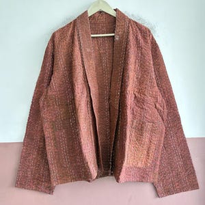 Puede incluir: Una chaqueta de manga larga abierta por delante en un tono marrón rojizo apagado, con una superficie texturizada y patrones de costura blanca visibles. La prenda tiene un cuello ancho, mangas rectas y dos grandes bolsillos de parche frontales.