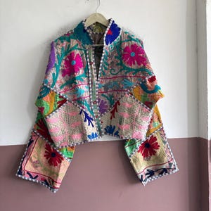 以下が含まれることがあります： 鮮やかなマルチカラーの刺繍が施されたキルティング生地のクロップドジャケット。長袖でスタンドカラーのデザインとなっており、前開き部分には明るい色のトリミングが施されています。ベースの生地はライトベージュです。