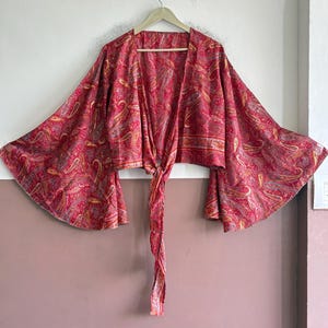 May include: A cropped wrap top featuring long, wide bell sleeves and a tie-front closure. The garment is made of a lightweight fabric with an intricate red, pink, and gold paisley pattern throughout, hanging on a light-colored plastic hanger.