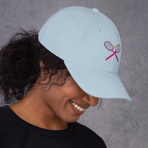 May include: Pale blue baseball cap with a pink tennis racket and ball emblem. The cap has a curved peak and a traditional six-panel design. The embroidered tennis graphic adds a sporty element to this casual accessory.
