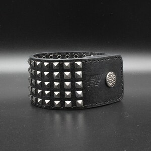 May include: A black leather cuff bracelet featuring four horizontal rows of silver-toned pyramid studs. The bracelet includes a silver-toned metal snap button closure embossed with a lightning bolt symbol and visible stitching along the edges.