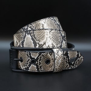 May include: A belt featuring a snakeskin pattern in shades of beige, brown, and black. The belt includes a matte black buckle and matching black hardware, with the word LOWLIFE embossed on the buckle and a small logo patch on the strap.