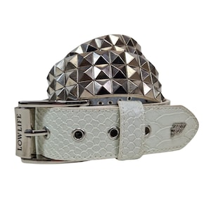 May include: White belt with a textured pattern and silver pyramid studs. The belt has a silver buckle and the text "LOWLIFE" on the buckle. The belt also has a silver emblem on the end.