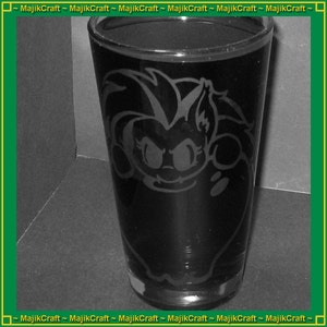 May include: A black pint glass with a cartoon character etched on the front. The glass has a wide base and tapers slightly towards the top. The etched design is a light color, contrasting against the dark glass. The glass is empty.