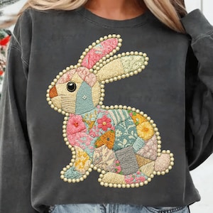 May include: Dark gray sweatshirt featuring a colorful patchwork bunny design. The bunny is adorned with floral and geometric patterns in shades of pink, blue, yellow, and orange, outlined with a beaded border. The sweatshirt has long sleeves and a crew neck.