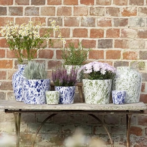 Op de afbeelding: Een verzameling keramische bloempotten met een gespikkeld ontwerp in verschillende maten en kleuren, waaronder blauw, groen en wit. De potten zijn gevuld met een assortiment planten en bloemen, tegen een bakstenen muur.