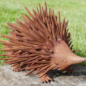 Puede incluir: Escultura de erizo de metal oxidado, con una cabeza detallada y un cuerpo cubierto de púas. La escultura es de color marrón rojizo y se encuentra sobre una superficie de madera. Decoración de jardín.