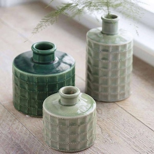 Puede incluir: Tres jarrones de cerámica verdes de diferentes tamaños. Presentan un patrón texturizado en forma de cuadrícula y un cuello estrecho. Los jarrones están colocados sobre una superficie de madera clara, con una ramita verde en el fondo.