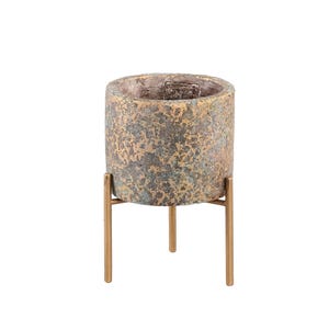 Puede incluir: Una maceta cilíndrica con un acabado texturizado y moteado en tonos bronce, cobre y gris oscuro. La maceta está sostenida por un soporte de metal dorado delgado de tres patas que eleva el recipiente, proporcionando una base estable.