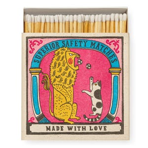 Pode incluir: Uma caixa de fósforos de madeira com pontas brancas. A caixa apresenta um fundo rosa com um leão amarelo e um gato malhado preto e branco a brincar com pequenas bolas. O texto diz SUPERIOR SAFETY MATCHES e MADE WITH LOVE.
