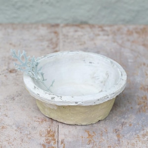 May include: A small, round, shallow bowl with a textured, pale yellow exterior and a thick, uneven white painted rim. A single, delicate, light-coloured, fern-like botanical sprig rests inside the bowl, extending over the edge of the rim.