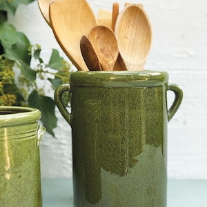 May include: A green ceramic utensil holder with two handles, filled with various wooden spoons and spatulas. The holder has a textured surface and a glossy finish. The kitchenware is set against a white background.