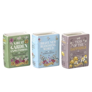 May include: Three ceramic book-shaped vases. One is green with "Great Garden Expectations" text, another is light blue with "Brave New Blooming World", and the third is purple with "Tess of the Daffodils". Each has floral designs.