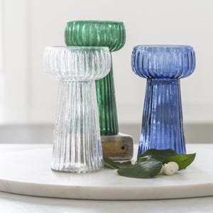 May include: Three glass candle holders in clear, green, and blue. Each has a ribbed design and a wide, shallow bowl shape. The clear holder is in the front, with the green and blue ones behind it. The candle holders are on a white marble surface.