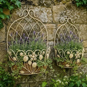 May include: Two cream-coloured metal wall planters with a lattice arch design and decorative floral and leaf accents. Each planter holds green foliage and purple lavender flowers, featuring a curved top hook for mounting on a stone wall surface.