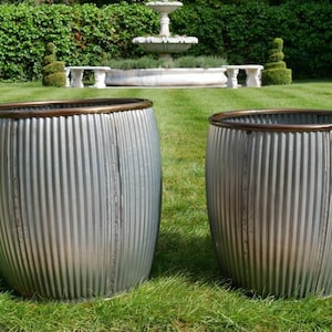 Peut inclure: Deux jardinières en métal galvanisé avec un design nervuré et des bords de couleur bronze. Les jardinières sont cylindriques et reposent sur une pelouse. L'arrière-plan présente une fontaine et des haies taillées, suggérant une décoration extérieure.