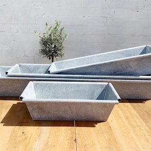 Puede incluir: Un conjunto de jardineras rectangulares de metal galvanizado de varios tamaños. Las jardineras tienen un aspecto gris desgastado con detalles negros en las esquinas. Un pequeño olivo es visible en el fondo, sugiriendo un tema de jardín.