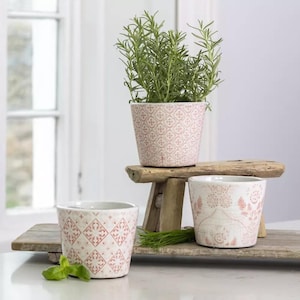 May include: Three decorative ceramic flower pots with a pink and white pattern. One pot holds a rosemary plant. The pots are on a wooden stand and a wooden tray. The pots have a vintage, farmhouse style.
