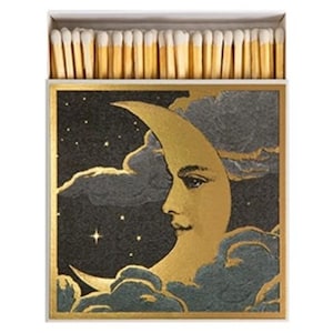 May include: A rectangular matchbox filled with wooden matches featuring white tips. The front of the box displays a gold crescent moon with a human face profile, set against a dark charcoal sky with gold stars and textured gray clouds.