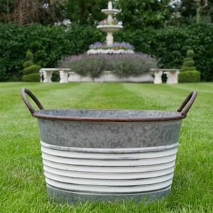May include: A galvanized metal tub with a brown rim and handles, set on a grassy lawn. The tub has horizontal white stripes around the exterior. A fountain and manicured garden are in the background.