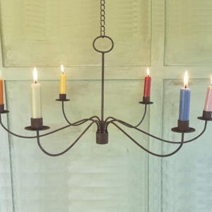 May include: A metal chandelier with six lit candles in different colours: orange, cream, yellow, red, blue, and pink. The chandelier hangs from a chain and has a central stem with curved arms holding the candle holders.