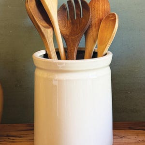 May include: A cylindrical white ceramic utensil crock filled with a collection of wooden kitchen tools, including various spoons and a salad fork. The container has a smooth, glossy finish and a thick rim, holding five distinct wooden utensils.