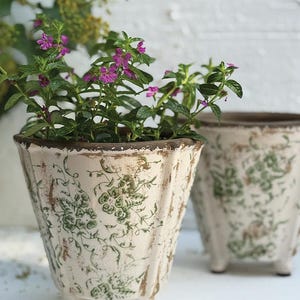 May include: Two off-white, distressed ceramic planters with green floral patterns. One planter is filled with green plants and small purple flowers. The planters have a vintage, rustic aesthetic and are elevated on small feet.