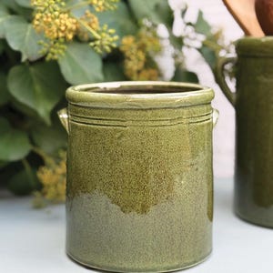 Può includere: Un portautensili in ceramica verde con finitura maculata. Il contenitore cilindrico ha due piccole maniglie e una superficie strutturata. L'utensile da cucina è di colore verde oliva tenue ed è adatto per riporre gli utensili da cucina.