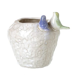 May include: A textured, off-white ceramic vase with a small opening. Two decorative birds perch on the rim: one blue with white speckles, the other light green. The vase has a rough, organic texture.