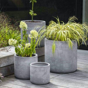 Puede incluir: Un conjunto de cuatro macetas de hormigón gris de varios tamaños, dispuestas sobre una terraza de madera. Las macetas contienen una variedad de plantas con follaje verde y amarillo, incluidas plantas con flores. Las macetas tienen un diseño cilíndrico simple.