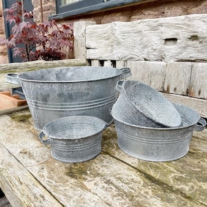 Puede incluir: Un conjunto de cuatro jardineras de metal galvanizado de diferentes tamaños. Las jardineras tienen un acabado envejecido y desgastado, con pequeñas asas laterales. La jardinera más grande mide unos 46 cm de diámetro, la más pequeña unos 15 cm.