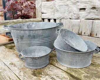 Puede incluir: Un conjunto de cuatro jardineras de metal galvanizado de diferentes tamaños. Las jardineras tienen un acabado envejecido y desgastado, con pequeñas asas laterales. La jardinera más grande mide unos 46 cm de diámetro, la más pequeña unos 15 cm.
