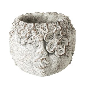 May include: A distressed, off-white ceramic planter in the shape of a face, with floral detailing around the top and over one eye. The lips are pursed. Ideal for small plants or flowers, this planter has a rustic aesthetic.