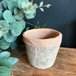 May include: A small, weathered terracotta pot with a textured, light-colored exterior and a faded orange rim. The pot is empty and sits on a wooden surface, with green foliage in the background.