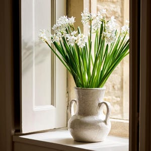 May include: A grey ceramic vase with two handles displays a bunch of white flowers with green stems. The vase is on a white windowsill. The background features a stone wall and a white window, creating a natural aesthetic.