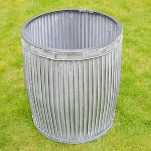 May include: A weathered, galvanized metal planter with a ribbed design. The cylindrical container has a dark rim and sits on a grassy surface. This outdoor planter is suitable for various plants and flowers.