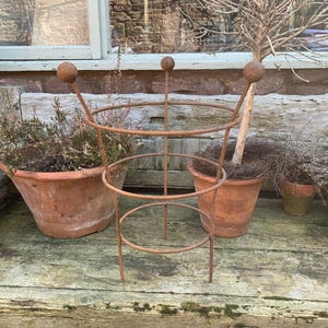 Peut inclure: Un support de plante en métal brun rouillé avec trois niveaux circulaires et deux boules décoratives. Le support est entouré de pots en terre cuite avec des plantes, posé sur une surface en bois vieilli.