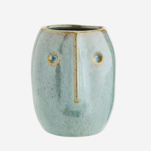 May include: A light blue ceramic vase with a face design. The vase has a rounded, oval shape with a flat top. The face features two circular eye indents, a vertical line for the nose, and a small mouth. The vase has a speckled, glazed finish.